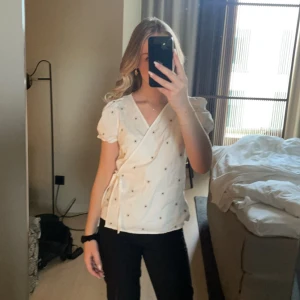 Vit omlottblus med blommönster - Säljer en söt vit omlottblus med små blommor. Blusen har korta ärmar och en knytning i sidan. Perfekt för vår och sommar!