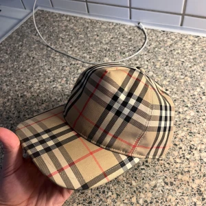 Burberry keps  - Snygg rutig keps från Burberry i klassiskt beige, svart och rött mönster. Kepsen har justerbar rem baktill och en broderad Burberry-logga. Perfekt för en stilren look. Sälj pgd av ingen användning. 9/10, helt ny. Hör av er priv!