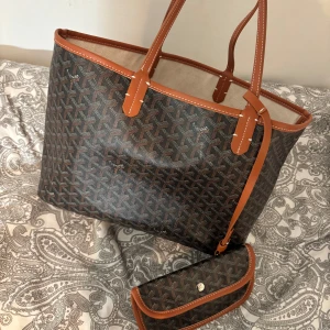 Mönstrad shoppingväska med matchande plånbok - Snygg shoppingväska med ett klassiskt mönster och matchande plånbok. Väskan har bruna handtag och detaljer, perfekt för en stilren look. Plånboken har en knappstängning och samma mönster som väskan.