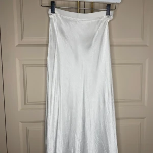 Vit långkjol från Divided - Elegant vit långkjol från Divided i ett glansigt material. Kjolen har en enkel och stilren design som passar perfekt för olika tillfällen. Den är lätt att matcha med olika toppar och accessoarer.