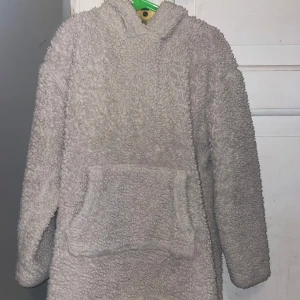 Grå teddyjacka från Lindex - Mysig grå teddyjacka från Lindex med huva och stor ficka framtill. Perfekt för kyliga dagar när man vill hålla sig varm och bekväm. Jackan har långa ärmar och en avslappnad passform.