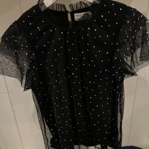 Svart prickig blus från Holly & Whyte - Snygg svart blus från Holly & Whyte( köpte den på Lindex) med vita prickar och volangdetaljer. Blusen har korta ärmar och en transparent look som ger en elegant touch. Perfekt för att piffa upp din outfit! (Använt 1 gång)