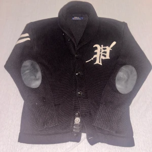 Svart stickad kofta från Polo Ralph Lauren - Säljer en svart stickad kofta från Polo Ralph Lauren med knappar och ett stort 'P' broderat på bröstet. Koftan har ribbade muddar och förstärkta armbågar i ett kontrasterande material. Perfekt för en stilren look. Men taggen undet vid höfterna fattas. Annars inget skada