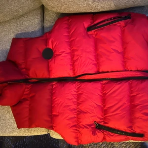Röd dunväst från Hugo Boss - Snygg röd dunväst från Hugo Boss med dragkedja och praktiska fickor. Perfekt för kyligare dagar och ger en stilren look. Västen har en svart insida med Hugo Boss-logga. Ny pris e runt 2500-3000