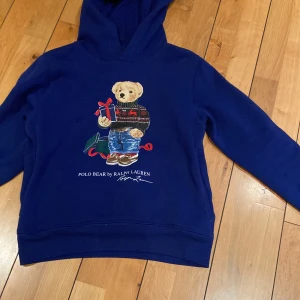 Blå hoodie från Ralph Lauren med nallemotiv - Säljer en blå hoodie från Ralph Lauren med ett  nallemotiv på framsidan. Nallen har en jultröja och håller i en present. Perfekt för en mysig och stilren look. 🐻🎁