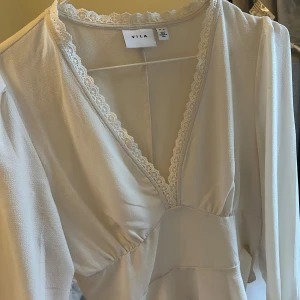 Vit tröja  från Vila - Elegant vit tröja från Vila med v-ringad spetsdetalj. Tröjan har långa ärmar och en mjuk, flödande design som ger en romantisk känsla. Perfekt för speciella tillfällen.