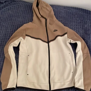 Nike tech fleece  - Säljer en snygg beige och vit hoodie från Nike med dragkedja och huva. Jackan har långa ärmar och en stilren design med svarta detaljer. Perfekt för en avslappnad stil.