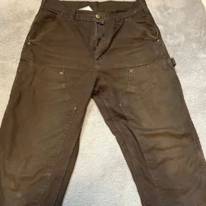  Carhartt byxor - Säljer ett par robusta bruna arbetsbyxor från Carhartt. Byxorna har flera praktiska fickor och är tillverkade i 100% bomull för hållbarhet och komfort. Perfekta för arbete eller fritid.