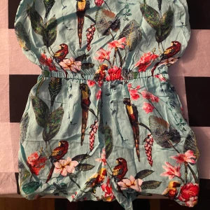 Blommig jumpsuit med papegojor - Snygg och färgglad jumpsuit med blommigt mönster och papegojor. Den har en elastisk midja och ärmlös design, perfekt för varma dagar. Den ljusgröna basen gör att de röda och rosa detaljerna verkligen poppar.