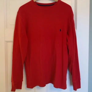 Röd långärmad tröja från Polo Ralph Lauren - Snygg röd långärmad tröja från Polo Ralph Lauren i 100% bomull. Den har en klassisk design med en liten broderad logotyp på bröstet. Perfekt för en stilren och avslappnad look.