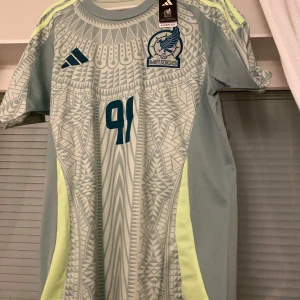 Mexico fotbollströja från Adidas - Säljer fotbollströja från Adidas med Mexikos landslagsmärke och nummer 91. Tröjan har korta ärmar och ett unikt mönster i ljusgrönt och grått. Perfekt för fotbollsfans!