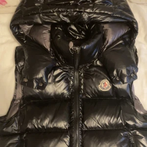 Svart dunväst från Moncler - Snygg svart dunväst från Moncler med glansig yta och dragkedja framtill. Västen har en hög krage och Moncler-logga på bröstet. Perfekt för kyliga dagar.
