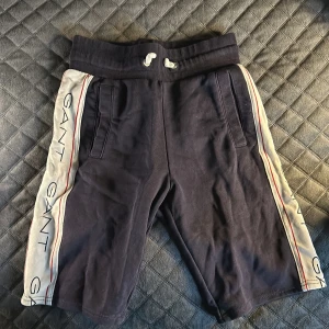 Mörkblå shorts från GANT - Snygga mörkblå shorts från GANT med vita sidopaneler och röd rand. De har snörning i midjan och fickor på sidorna. Perfekta för en avslappnad stil.