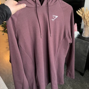 Lila/vinröd hoodie - Säljer en lila/vinröd hoodie från Gymshark. Perfekt för träning eller en avslappnad dag. Den har en diskret logga på bröstet och är tillverkad i ett mjukt material.