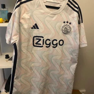 Vit Ajax fotbollströja - Snygg vit Ajax fotbollströja från Adidas med korta ärmar och ett unikt mönster. Tröjan har klubbens emblem och sponsorlogga på framsidan samt tre stjärnor ovanför emblemet. Perfekt för fans av laget!