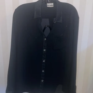 Svart blus från Lecomte - Elegant svart blus från Lecomte med knappar framtill och en diskret bröstficka med broderade tecken. Perfekt för en stilren look.