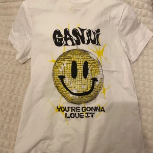 Vit t-shirt med smiley från Ganni - Säljer en vit t-shirt från Ganni med ett stort gult smileytryck och text. Kortärmad och i bomullsmaterial. Tvättas innan den skickas. Använd typ 3 gånger!!🥰 pris kan diskuteras. 