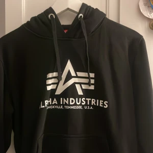 Svart hoodie från Alpha Industries - Snygg svart hoodie från Alpha Industries med deras ikoniska logga i vitt på bröstet. Tröjan har en klassisk känguruficka och justerbar huva med dragsko. Perfekt för en avslappnad stil.