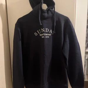 Svart hoodie från Sunday Sportswear - Säljer en svart hoodie från Sunday Sportswear med texten 'Sunday Sportswear Est. 1973' på bröstet. Den har en justerbar huva med dragsko och långa ärmar. Perfekt för en avslappnad stil.