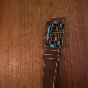 Brunt skinnbälte från J. Lindeberg - Snyggt brunt skinnbälte från J. Lindeberg med en unik spänne i geometrisk design. Tillverkat i Sverige av äkta läder, perfekt för att ge en stilren touch till din outfit.