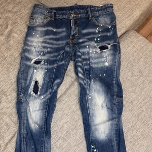 Blå jeans från Dsquared2 - Säljer ett par blå jeans från Dsquared2 med slitna detaljer och färgstänk. Jeansen har en klassisk femficksdesign och knappgylf. Säljs då jag bytt stil.