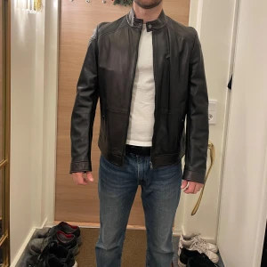 Svart skinnjacka från Boss - Snygg svart skinnjacka från Boss med dragkedja och ståkrage. Jackan har en stilren design med två fickor framtill och knappar vid ärmsluten. Perfekt för en cool och tidlös look. Originalpris: 4199 kr