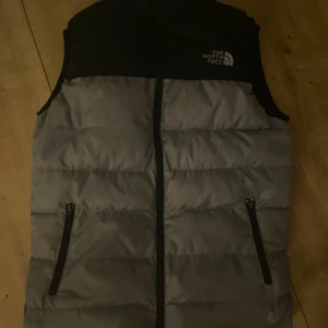 Grå och svart dunväst från The North Face äkta  - Säljer en snygg grå och svart dunväst från The North Face. Västen har en dragkedja framtill och två praktiska fickor med dragkedjor. Perfekt för kyligare dagar.