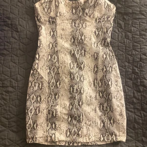 Ormmönstrad axelbandslös klänning - Trendig axelbandslös klänning med ormmönster i svart och beige. Klänningen har en dragkedja bak och en figurnära passform som framhäver silhuetten. Perfekt för en utekväll eller speciella tillfällen. Vadderad typ vid bysten så man inte behöver ha bh!