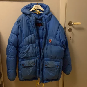 Blå dunjacka från Fjällräven - Säljer en blå dunjacka från Fjällräven med huva och praktiska fickor framtill. Jackan har en dragkedja och knappar för extra skydd mot kyla. Perfekt för kalla vinterdagar. Lägg dock märke till att fickorna är lite missfärgade. Jag som är 160 cm lång och väger 44 kg upplever den som för stor.💕