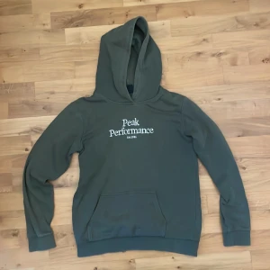 Grön hoodie från Peak Performance - Säljer en stilren grön hoodie från Peak Performance. Den har en klassisk design med en stor ficka framtill och en bekväm huva. Perfekt för en avslappnad look. Storlek 170. 9/10 i skick