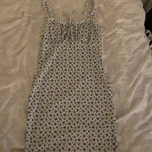 Blommig klänning med knytband - Charmig klänning med blå blommor på vit bakgrund. Den har smala axelband och ett sött knytband framtill. Perfekt för sommarens varma dagar.