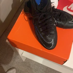 Svarta fotbollsskor från Nike - Säljer ett par svarta fotbollsskor från Nike med blå och vita detaljer. Skorna har snörning och dobbar för bra grepp på planen. Perfekta för fotbollsträning och matcher.