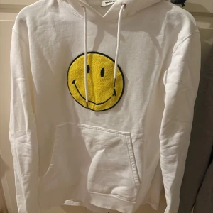 Vit hoodie från Sandro med smiley - Säljer en vit hoodie från Sandro med en stor gul smiley på framsidan. Hoodien har en klassisk design med dragsko i huvan och en praktisk magficka. Perfekt för en avslappnad stil! Använd en gång