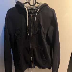 Mörkblå hoodie från GANT - Säljer en snygg mörkblå hoodie från GANT med dragkedja och justerbar huva. Tröjan har ett broderat GANT-märke på framsidan och praktiska fickor. Perfekt för en avslappnad stil.