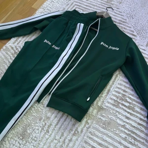 Grön träningsset från Palm Angels - Snyggt grönt träningsset från Palm Angels med vita ränder längs ärmar och ben. Jackan har dragkedja och byxorna har resår i midjan. Perfekt för en sportig look.