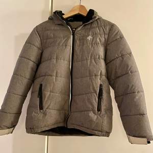 Snygg grå dunjacka från Cross Sportswear med huva och dragkedja. Jackan har två praktiska fickor med dragkedjor och är perfekt för kyliga dagar. Den quiltade designen ger en modern look.
