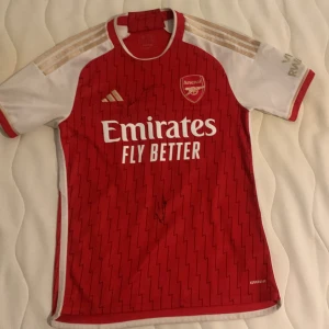 Singnerad Röd Arsenal fotbollströja med Martinelli 11 - Säljer en röd Arsenal fotbollströja från Adidas med Martinelli och nummer 11 på ryggen. Tröjan har korta ärmar och ett subtilt mönster. Den är prydd med 'Emirates Fly Better' på framsidan och har klubbens emblem på bröstet. Perfekt för fans som vill visa sitt stöd! Tröjan är även singnerad av Arsenal spelarna Trossard och Martinelli