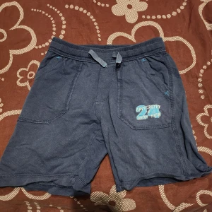 Mörkblå shorts med tryck - Säljer ett par mörkblå shorts med tryck på ena benet. De har en bekväm passform med snörning i midjan och praktiska fickor framtill. Perfekta för en avslappnad stil.