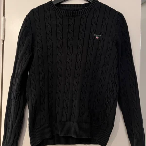 Svart kabelstickad tröja från GANT - Snygg svart kabelstickad tröja från GANT i premium bomull. Tröjan har en klassisk rund halsringning och ribbade muddar vid ärmslut och nederkant. Perfekt för en stilren look.