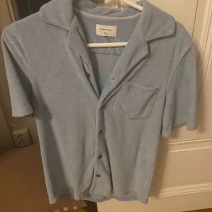 Ljusblå frottéskjorta från Studio Total - Säljer en ljusblå frottéskjorta från Studio Total. Skjortan har korta ärmar, en bröstficka och knappar framtill. Perfekt för en avslappnad stil.