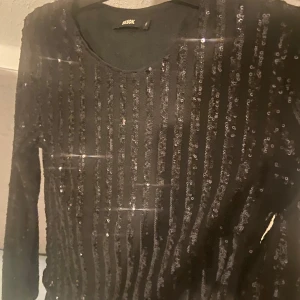 Svart glittrig tröja från ASOS - Snygg svart tröja från ASOS med glittriga paljetter i ett randigt mönster. Perfekt för en kväll ute eller festliga tillfällen. Tröjan har långa ärmar och en rund halsringning.