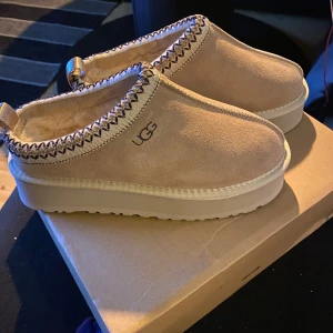 Beige mockaskor från UGG - Säljer ett par snygga beige mockaskor från UGG med fluffigt foder och dekorativ söm runt kanten. Skorna har en robust sula och är perfekta för kyligare dagar. De är både stiliga och bekväma.