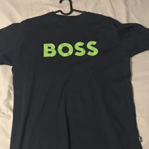 Mörkblå t-shirt från Boss - Snygg mörkblå t-shirt från Boss med stort grönt tryck på bröstet. Perfekt för en avslappnad stil. Kortärmad och tillverkad i mjukt material för extra komfort.