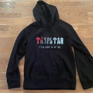 Svart hoodie från Trapstar - Säljer en svart hoodie från Trapstar med texten 'Trapstar It's a Secret' i rött och vitt på framsidan. Hoodien har en klassisk design med en stor ficka framtill och en justerbar huva. Perfekt för en avslappnad stil.