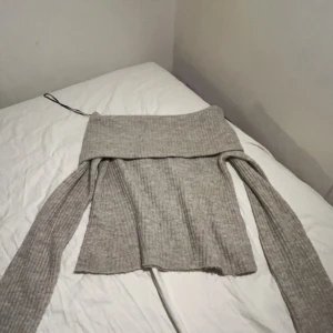 Grå ribbad offshoulder tröja från H&M - Säljer en stilren grå ribbad offshoulder tröja från H&M. Tröjan har långa ärmar och en bred krage som ger en avslappnad look. Perfekt för kyligare dagar. Använd en gång 