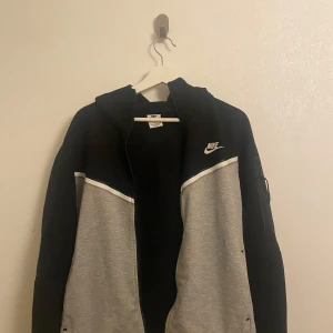 Svart och grå hoodie från Nike - Säljer en snygg svart och grå hoodie från Nike med dragkedja och huva. Jackan har en klassisk design och är perfekt för en sportig look. Den köptes för 1,5 år sedan och har använts vissa gånger men inget fel på den!