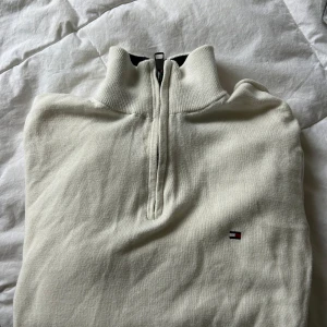 Vit tröja med dragkedja från Tommy Hilfiger - Säljer en stilren vit tröja från Tommy Hilfiger med halv dragkedja och broderad logotyp på bröstet. Tröjan har långa ärmar och ribbade muddar vid ärmslut och nederkant. 