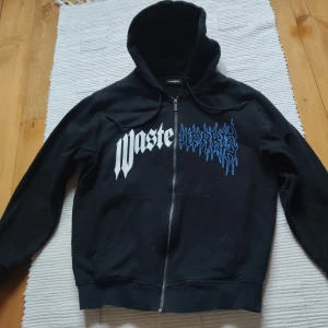 Svart hoodie från Wasted Paris, st M - Svart hoodie från Wasted Paris med tryck i vitt och blått på framsidan. Hoodien har en dragkedja och en klassisk passform med huva. Tillverkad i tjock och varm bomull. Använt 1-2 gånger!