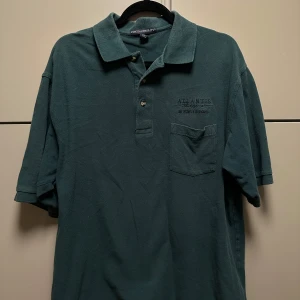 Grön pikétröja från Port Authority - Säljer en klassisk thriftad grön pikétröja från Port Authority. Tröjan har en krage med knappar och en bröstficka. Perfekt för en avslappnad stil. Klippt snyggt så den är bra längd. 