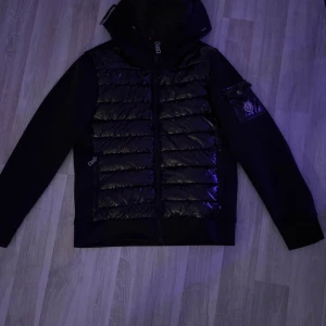 Svart jacka från Moncler - Snygg svart jacka från Moncler med quiltad framsida och huva. Jackan har en dragkedja och en ficka med knappdetalj på ärmen. Perfekt för kyligare dagar.